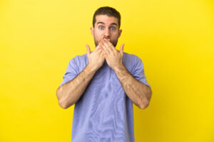 Handsome blonde man over isolated yellow background covering mouth with hands